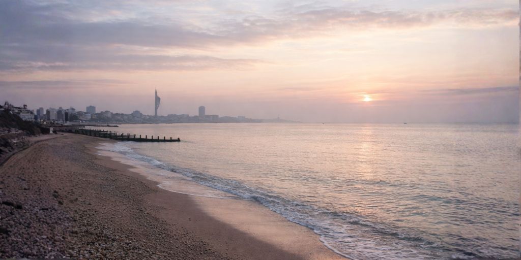 Sunrise over a quiet beach with calm sea and distant city skyline.